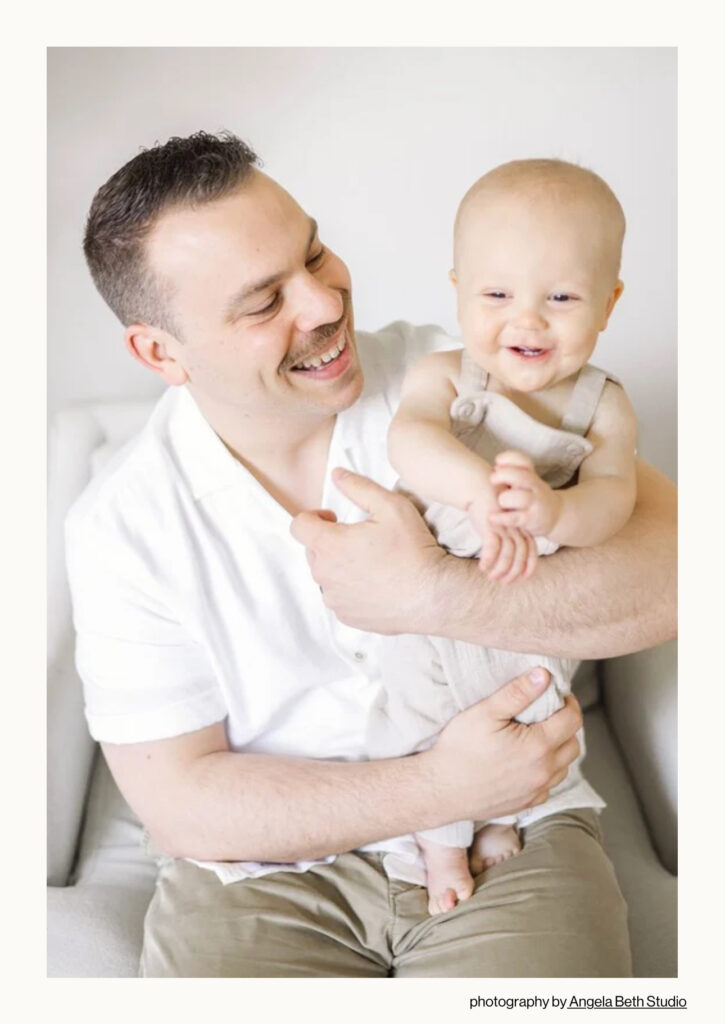 Father holding smiling baby boy during an in-home first birthday session in Northern NJ, captured by Angela Beth Studio. Featured in Feels Like Family Magazine to honor the quiet beauty of fatherhood through natural, lifestyle photography.