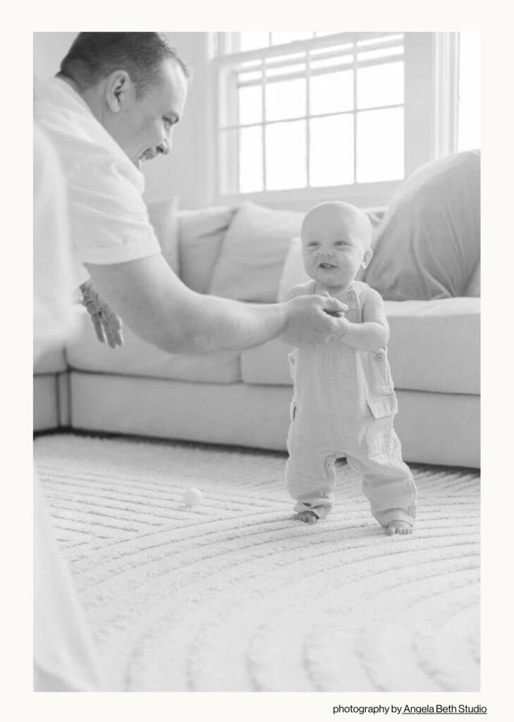 Black and white photo of a father helping his baby stand and take steps during a documentary-style milestone session in Wayne NJ. Photographed by Angela Beth Studio and nationally featured for its tender portrayal of fatherhood.