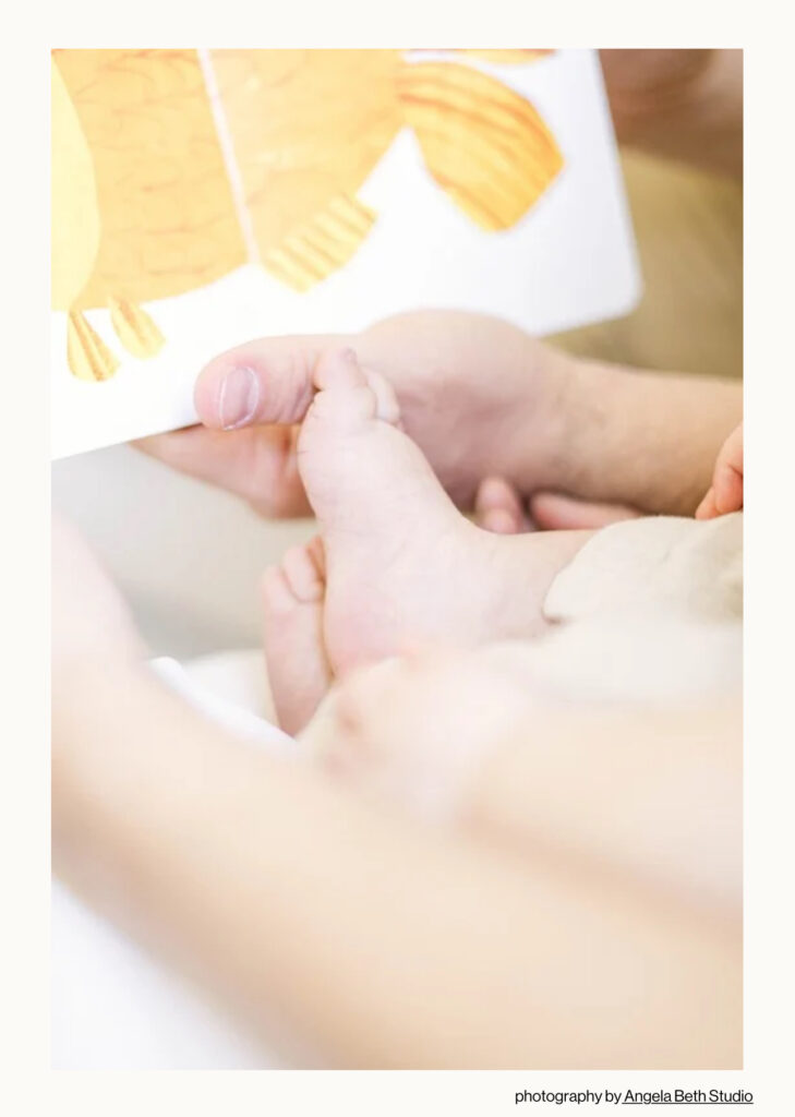 Close-up of a father holding his baby’s feet while reading a picture book during a lifestyle milestone session in Northern NJ. Captured by Angela Beth Studio and featured in Feels Like Family Magazine to celebrate quiet fatherhood moments.