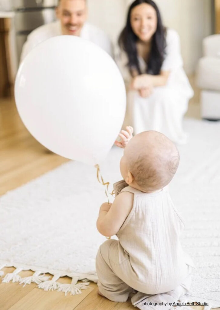 Baby playing with a white balloon while parents watch in the background during a joyful in-home first birthday session in Bergen County NJ. Photographed by Angela Beth Studio and recognized in Feels Like Family Magazine’s Story Worthy feature.